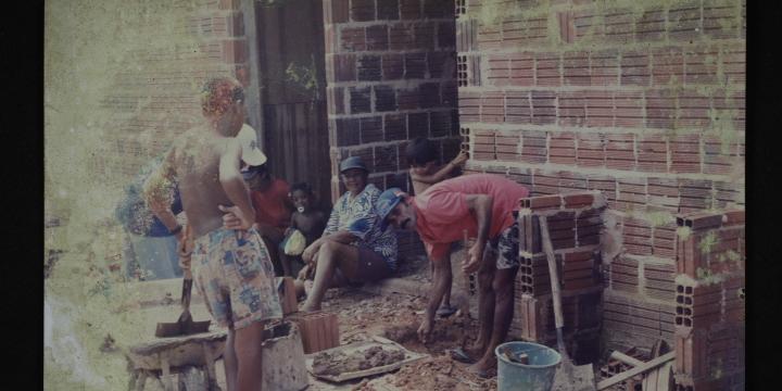 Foto antiga, manchada, de um mutirão de construção. O registro mostra homens e mulheres trabalhando com tijolos, materiais de construção e terra na frente de uma parede de tijolos aparentes.