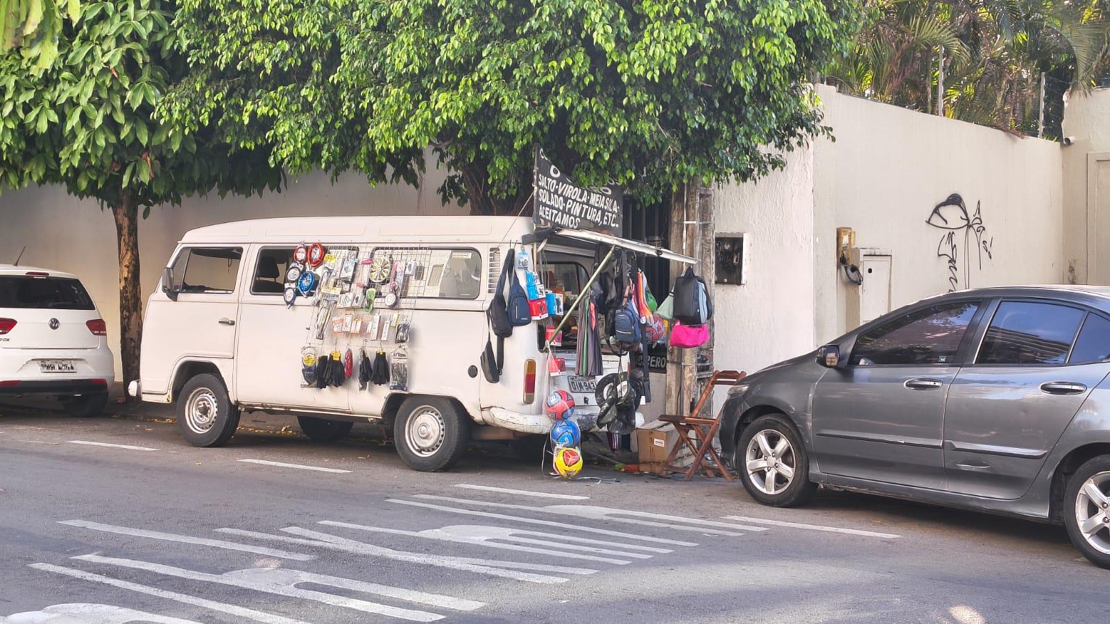 Imagem mostra veículo kombi estacionado e servindo de loja com produtos para vender pendurados para o lado de fora.